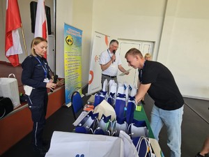 Zdjęcie Kolorowe. Jeden z uczestników eliminacji Ogólnopolskiego Młodzieżowego Turnieju Motoryzacyjnego pochyla się nad stołem, gdzie znajdują się nagrody. Na wprost niego stoi policjantka ruchu drogowego, która trzyma w dłoni mikrofon. W tle mężczyzna, który w jednej dłoni trzyma kartkę papieru w drugiej mikrofon do którego mówi.