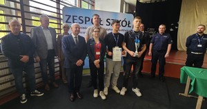 Zdjęcie kolorowe. Trzyosobowa drużyna uczestników turnieju wraz z opiekunem pozują do zdjęcia. Jeden z uczniów trzyma w dłoni puchar drugi dyplom. Po ich lewej stronie stoi dwóch policjantów, po prawej stoją organizatorzy turnieju.