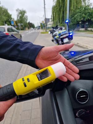 Zdjęcie kolorowe. Urządzenie do badania stanu trzeźwości trzymane w dłoni, świeci na czerwono. W tle policyjny motocykl.
