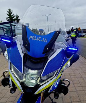 Zdjęcie kolorowe. Na pierwszym planie policyjny motocykl z włączonymi światłami błyskowymi. W tle policjant - motocyklista rozmawiający z zatrzymanym do kontroli kierowcą.