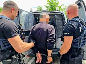 Zdjęcie kolorowe. Dwóch policjantów przytrzymujących zatrzymanego na tle otwartych drzwi radiowozu służącego do przewożenia osób zatrzymanych.