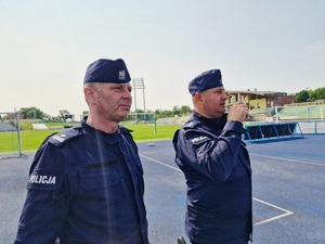 Zdjęcie kolorowe. Dwóch funkcjonariuszy dowodzących ćwiczeniami w umundurowaniu ćwiczebnym, na tle murawy stadionu. Jeden z policjantów rozmawia przez krótkofalówkę.