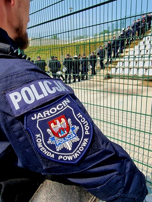 Zdjęcie kolorowe. Na pierwszym planie ramie policjanta z naszywką Jarocin Komenda Powiatowa Policji. W tle rząd policjantów ćwiczących na pustych trybunach stadionu.