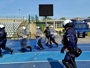 Zdjęcie kolorowe. Policjanci w umundurowaniu ćwiczebnym wyposażeni w hełmy ochronne i tarcze wbiegają na stadion. W tle specjalistyczny samochód policyjny wyposażony w armatki wodne.