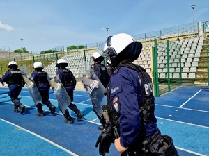 Zdjęcie kolorowe. Policjanci w umundurowaniu ćwiczebnym wyposażeni w hełmy ochronne i tarcze wbiegają na stadion. W tle puste trybuny stadionu.