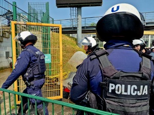 Zdjęcie kolorowe. Policjanci w umundurowaniu ćwiczebnym wyposażeni w hełmy ochronne i tarcze wchodzą na teren pustego sektora kibiców przyjezdnych. 
Na pierwszym planie funkcjonariusz odwrócony tyłem, na umundurowaniu duży napis POLICJA.