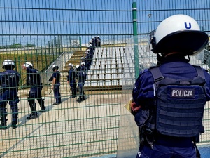 Zdjęcie kolorowe. Ustawieni w rzędzie policjanci w umundurowaniu ćwiczebnym wyposażeni w hełmy ochronne oraz pałki ćwiczą na teren pustego sektora kibiców przyjezdnych. 
Na pierwszym planie funkcjonariusz odwrócony tyłem, przed sobą trzyma tarczę.