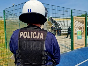 Zdjęcie kolorowe. Policjanci w umundurowaniu ćwiczebnym, stojący w rzędzie, wyposażeni w hełmy ochronne oraz pałki ćwiczą na terenie pustego sektora kibiców. Na pierwszym planie funkcjonariusz stojący tyłem, przed sektorem kibiców.