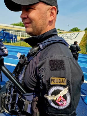 Zdjęcie kolorowe. Na pierwszym planie policjant, który na ramieniu ma tarczę z napisem - Grupa Realizacyjna Policja Kalisza
