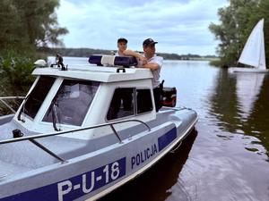 Zdjęcie kolorowe. Policjanci posterunku wodnego podczas służby na łodzi. Na łodzi napisy P-U-18 Policja. W tle na zbiorniku wodnym żaglówka.