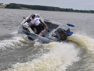 Zdjęcie kolorowe. Policjant posterunku wodnego stojący na płynącej łodzi patrolowej. W tle tereny leśne.