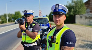 Zdjęcie kolorowe. Policjanci ruchu drogowego w umundurowaniu służbowym. Jeden z policjantów mierzy prędkość nadjeżdżających pojazdów. W tle droga oraz policyjny, oznakowany radiowóz zaparkowany za policjantami.