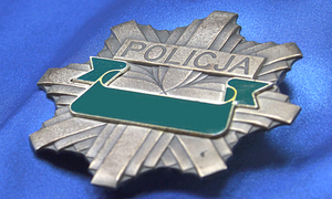 Policyjna odznaka koloru szarego, z zielona wstęga w środkowej części, położona na błękitnym tle.