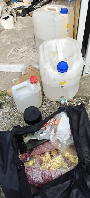 Zdjęcie kolorowe. Trzy białe pojemniki plastikowe z zawartością cieczy. Obok stoi otwarta czarna, sportowa torba w której znajdują się woreczki foliowe z zapakowanymi tabletkami koloru żółtego i czerwonego.