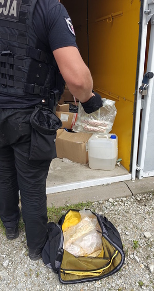 Zdjęcie kolorowe. Policjant trzymający w dłoni woreczki strunowe z zawartością różowych tabletek. Na ziemi leży czarna, sportowa torba z zawartością woreczków z tabletkami.