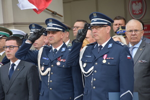 Zdjęcie Kolorowe. Dwaj policjanci w umundurowaniu galowym oddający honor. Obok, w tle stoją ludzie goście na uroczystości obchodów Święta Policji.