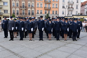Zdjęcie Kolorowe. Pododdział policjantów w umundurowaniu galowych stojący na Głównym Rynku w Kaliszu podczas uroczystości obchodów Święta Policji. Policjanci trzymają białe teczki z logo Policji.