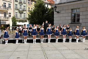 Zdjęcie Kolorowe. Na pierwszym planie dziewczynki w granatowych sukienkach i długich białych butach (mażoretki). W tle na podeście orkiestra. Po prawej stronie fragment ratusza. W tle po lewej widać kamienice.