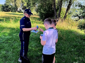 Zdjęcie kolorowe. Policjantka w umundurowaniu służbowym koloru granatowego rozdaje ulotki trzem chłopcom. W tle zbiornik wodny i drzewa.