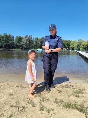Zdjęcie kolorowe. Policjantka w umundurowaniu służbowym koloru granatowego obok której stoi kilkuletnia dziewczynka ubrana w sukienkę. W tle zbiornik wodny.
