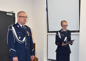 Zdjęcie kolorowe. Dwoje policjantów w umundurowaniu wyjściowym koloru granatowego, z białymi sznurami. Mężczyzna stoi po lewej stronie, kobieta po prawej.