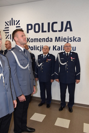 Zdjęcie kolorowe. Dwaj policjanci w umundurowaniu wyjściowym ze sznurem, czekający na wręczenie wyróżnień. Z prawej strony pod ścianą ustawieni policjanci - przełożeni, stojący pod napisem POLICJA Komenda Miejska Policji w Kaliszu.