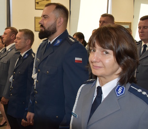 Zdjęcie kolorowe. Policjanci w umundurowaniu wyjściowym ze sznurem, stoją w szeregach oczekując na wręczenie aktów mianowania. Na pierwszym planie policjantka spoglądająca w aparat.