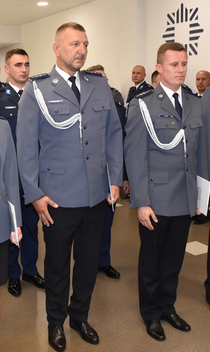 Zdjęcie kolorowe. Na  pierwszym planie dwóch policjantów w umundurowaniu wyjściowym z białym sznurem. Stoją w szeregu trzymając w dłoniach papierowe teczki koloru białego