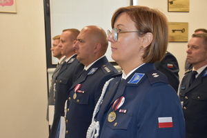 Zdjęcie kolorowe. Policjanci w umundurowaniu wyjściowym z białym sznurem stojący w szeregu. W tle widać część drugiego szeregu policjantów.