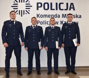 Zdjęcie kolorowe. Czterech policjantów w umundurowaniu koloru granatowego, pozują do zdjęcia na tle ściany, na której umieszczony jest napis Policja Komenda Miejska Policji w Kaliszu