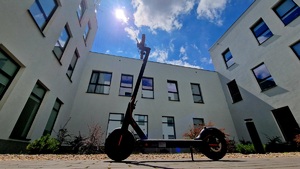 Zdjęcie kolorowe. Hulajnoga stojąca na chodniku. W tle okna budynku z trzech stron - z prawej, lewej i z tłu. U góry białe chmury i słońce na tle błękitnego nieba.