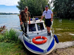 Zdjęcie kolorowe. Policyjna łódź przy zacumowana przy brzegu. Na jej pokładzie z prawej strony stoi policjant z lewej strony funkcjonariusz Państwowej Straży Łowieckiej