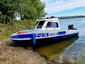 Zdjęcie kolorowe. Policyjna łódź patrolowa zacumowana przy brzegu. Z lewej burty napis P-U-18 Policja.