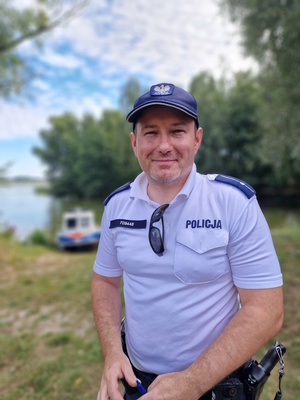 Zdjęcie kolorowe. Policjant z posterunku wodnego w umundurowaniu służbowym (biała koszulka z granatowym napisem Policja, granatowa czapka)