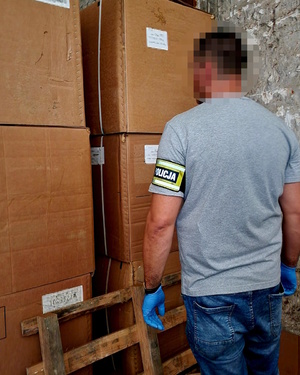 Zdjęcie kolorowe. Policjant nie umundurowany stojący tyłem.  Na lewym ramieniu ma opaskę z napisem policja. Funkcjonariusz stoi przed kartonami, które ułożone są jeden na drugim. O kartony na dole oparta jest drewniana paleta.