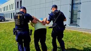 Zdjęci kolorowe. Dwóch policjantów prowadzi zatrzymanego, który na nadgarstkach ma założone kajdanki. Zdjęcie przed budynkiem komendy.