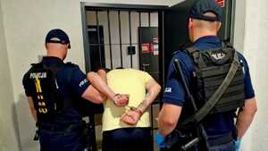 Zdjęci kolorowe. Dwóch policjantów stoi wraz z zatrzymanym przed kratą, która jest wejściem do pomieszczeń dla osób zatrzymanych w komendzie. Zatrzymany ma ręce z tyłu, na których założone są kajdanki.