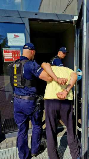 Zdjęci kolorowe. Jeden z policjantów wprowadza zatrzymanego do budynku komendy, drugi z funkcjonariuszy przytrzymuje otwarte drzwi.