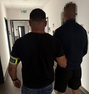 Zdjęcie kolorowe. Policjant nieumundurowany - na lewym ramieniu opaska z napisem &quot;Policja&quot; prawą ręką trzyma zatrzymanego mężczyznę. Obaj stoją tyłem. W tle korytarz.