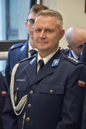 Zdjęcie kolorowe. Policjant w umundurowaniu wyjściowym ze sznurem.