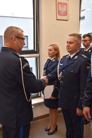 Zdjęcie kolorowe. Policjant w umundurowaniu wyjściowym ze sznurem, wręcza akty mianowani a policjantom stojącym w szeregu.