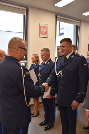 Zdjęcie kolorowe. Policjant w umundurowaniu wyjściowym ze sznurem, wręcza akty mianowani a policjantom stojącym w szeregu.