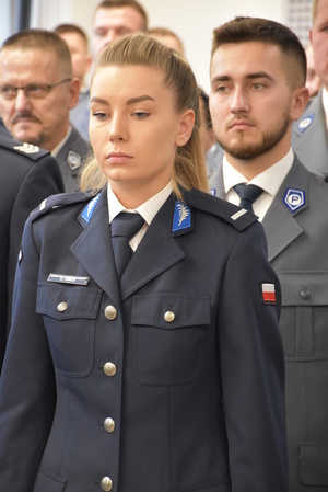 Zdjęcie kolorowe. Na pierwszym planie policjantka w umundurowaniu wyjściowym koloru granatowego. Za nią, w dalszych szeregach stoją policjanci ubrani w mundury wyjściowe, ale koloru stalowego.
