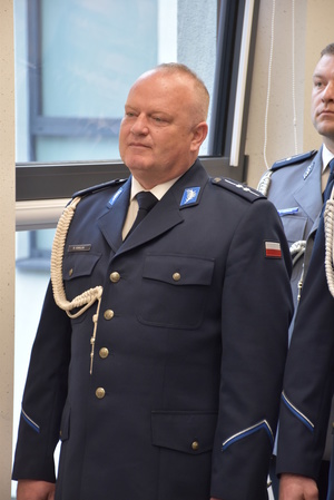 Zdjęcie kolorowe. Policjant w umundurowaniu wyjściowym koloru granatowego, ze sznurem. W tle okno.
