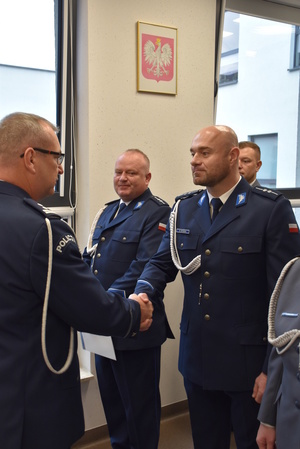 Zdjęcie kolorowe. Policjant w umundurowaniu wyjściowym ze sznurem, wręcza akty mianowani a policjantom stojącym w szeregu.