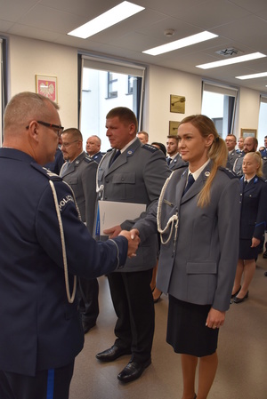 Zdjęcie kolorowe. Policjant w umundurowaniu wyjściowym ze sznurem, wręcza akty mianowani a policjantom stojącym w szeregu.
