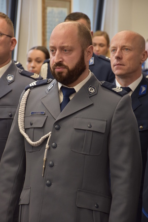 Zdjęcie kolorowe. Policjant w umundurowaniu wyjściowym ze sznurem. Mundur koloru stalowego. W tle, w kolejnych rzędach stoją policjanci.