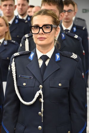 Zdjęcie kolorowe. Policjantka w umundurowaniu wyjściowym, ze sznurem. Mundur koloru granatowego. W tle widać policjantów stojących w szeregach.