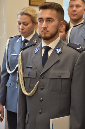 Zdjęcie kolorowe. Policjant w umundurowaniu wyjściowym koloru stalowego, ze sznurem. Za nim stoją w rzędach kolejni policjanci uczestniczący w uroczystości.
