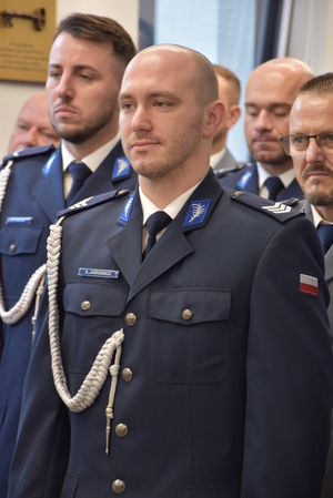 Zdjęcie kolorowe. Policjant w umundurowaniu wyjściowym koloru granatowego, ze sznurem. Za nim stoją w rzędach kolejni policjanci uczestniczący w uroczystości.